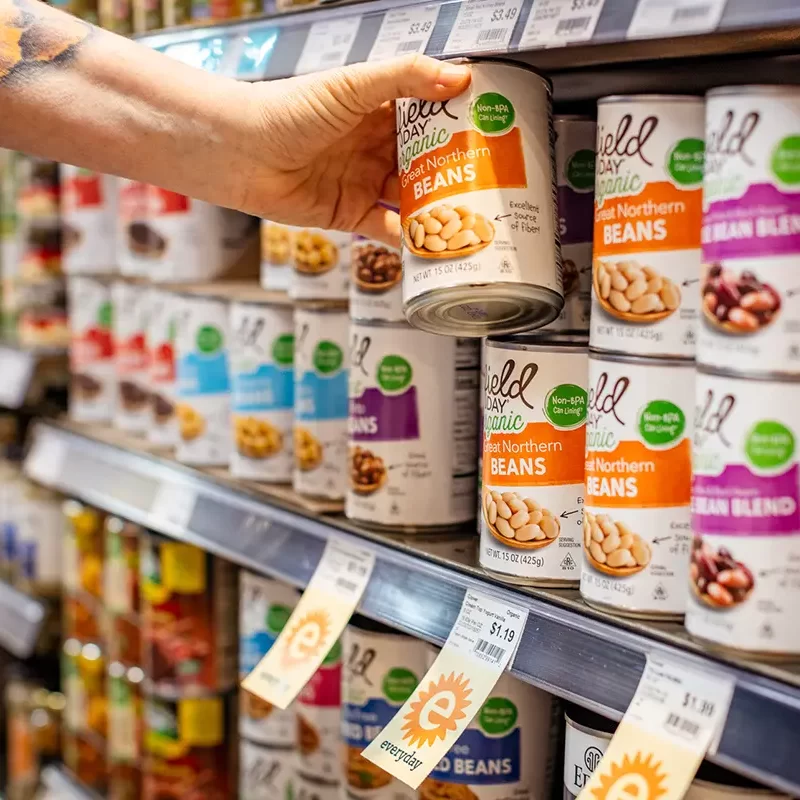 A hand reaching for a can of beans on a display rack.